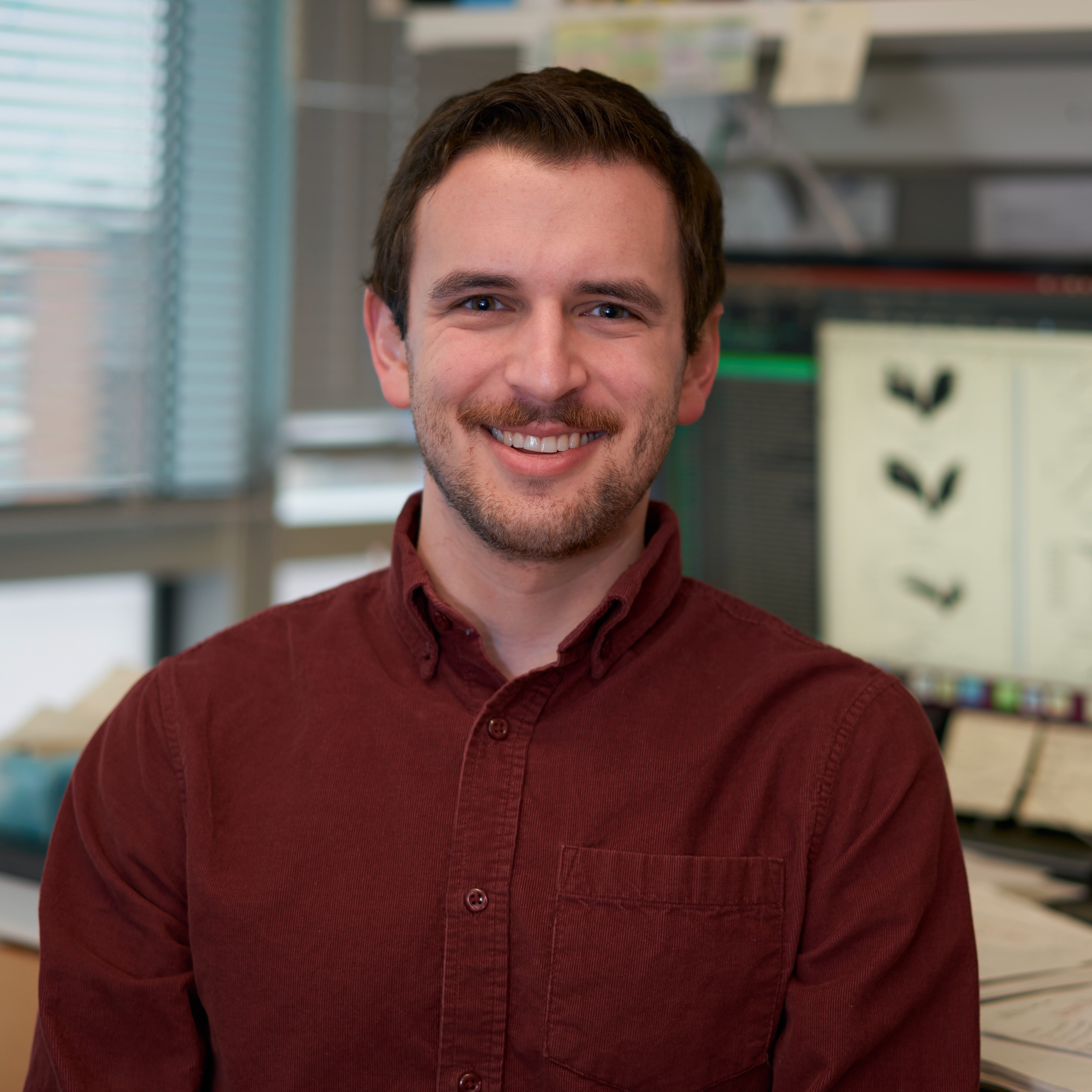 A brown-haired man with a short beard and mustache wearing a red corduroy shirt sitting in front of a comuputer screen showing volcano plots.