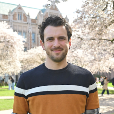 A man with curly brown hair, a short beard, mustache, and black hoop earrings wearing a multicolored striped sweater standing in front of cherry blossoms and an academic building.